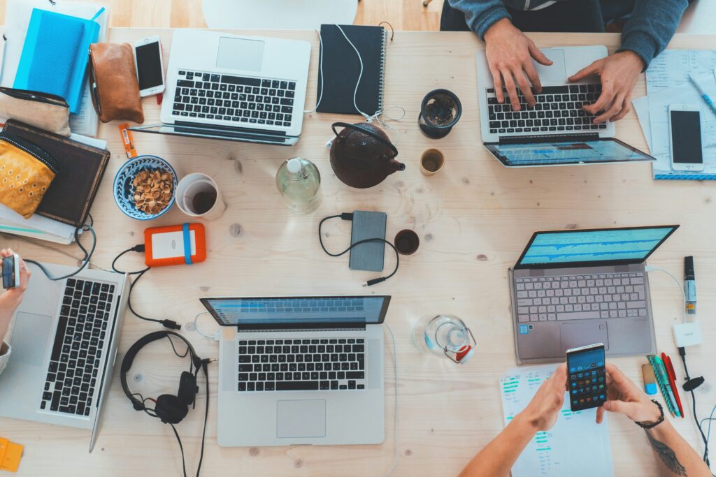 Four people collaborating at a cluttered wooden table with laptops, notebooks, snacks, headphones, and smartphones—perhaps brainstorming strategies for Brand Reputation Management—captured from an overhead perspective.